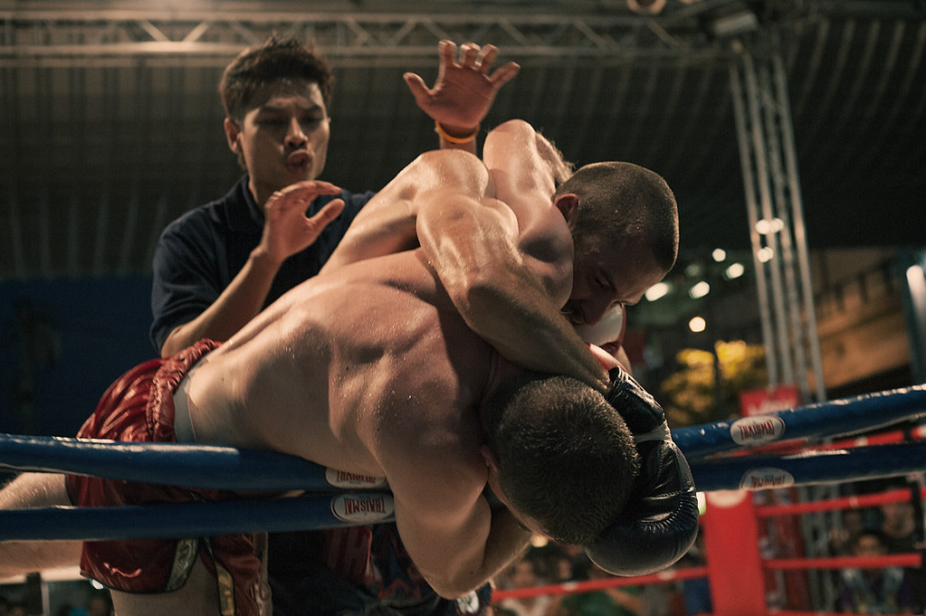 Two Muay Thai fighters clinch in the ring as the referee watches